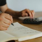 Hands writing calculations in a notebook beside a calculator
