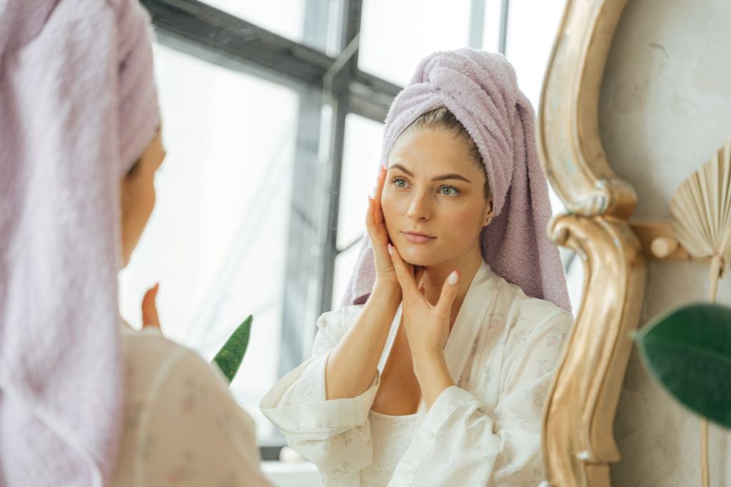 A woman in a robe, hair wrapped in a towel, touching her cheeks at a mirror by a bright window