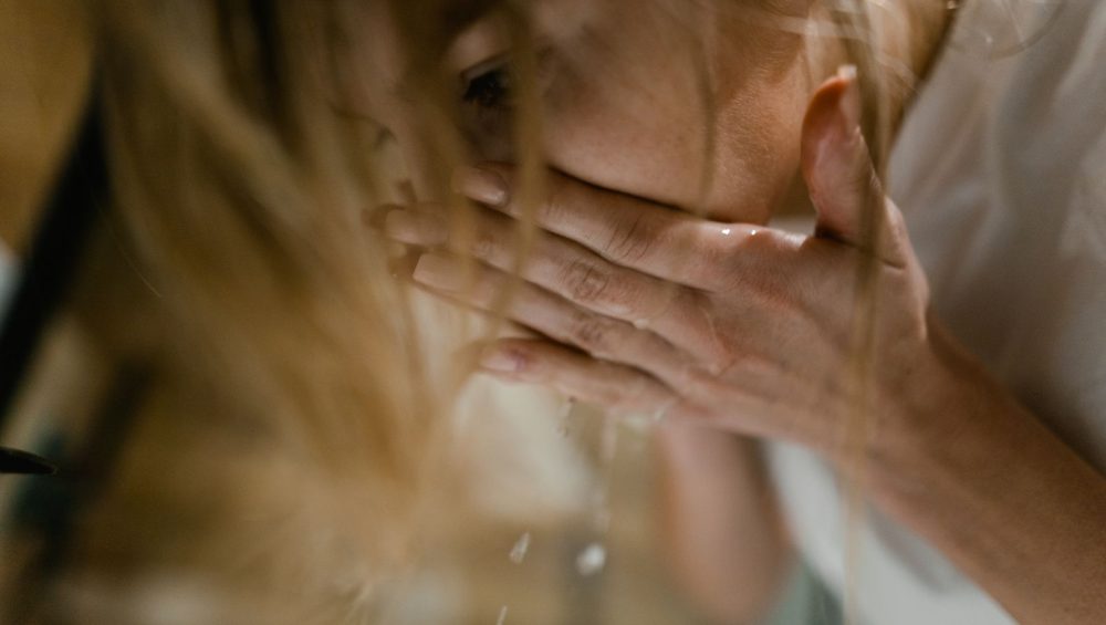 A woman rinsing her face with cupped hands over a basin, water dripping from her fingers in warm light