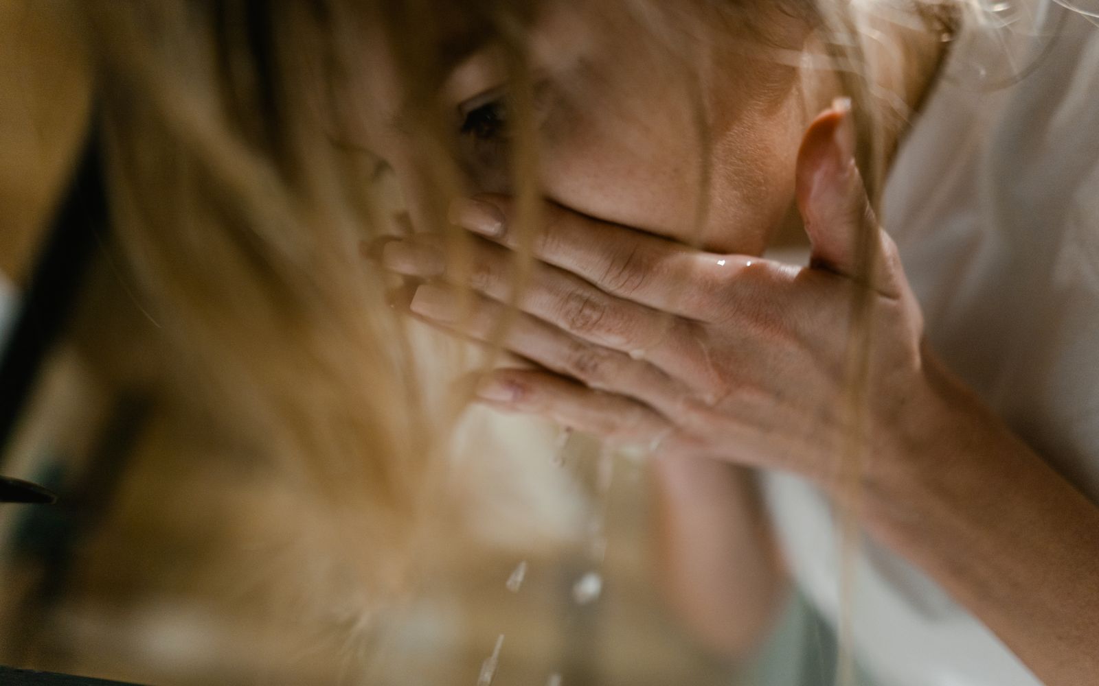 A woman rinsing her face with cupped hands over a basin, water dripping from her fingers in warm light