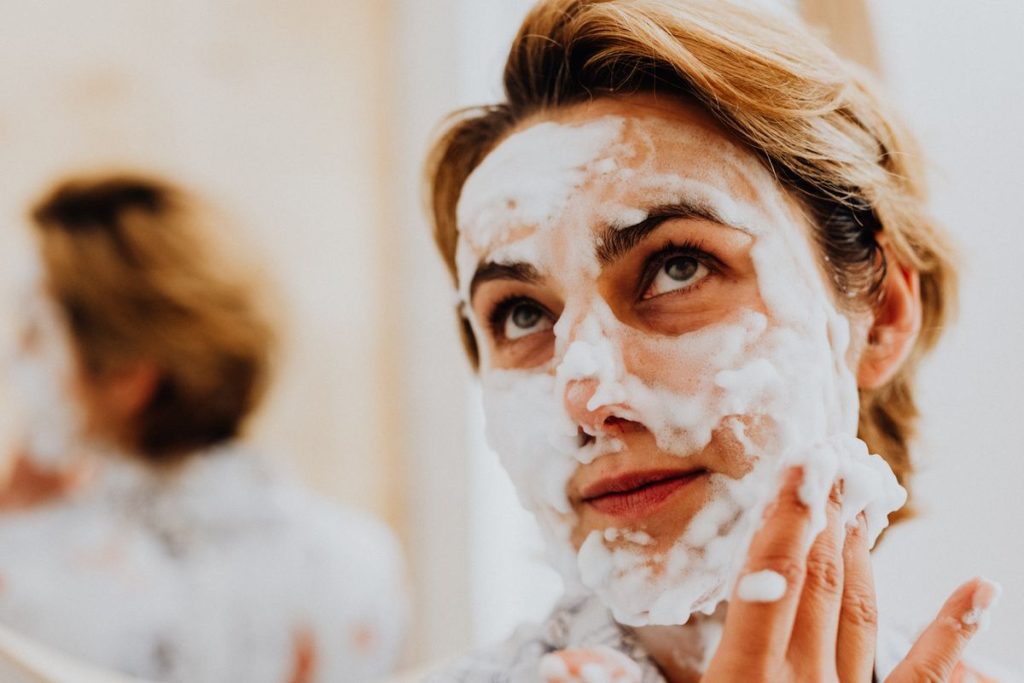 Close view of a woman with a cleanser lather across her face, her fingertips working it gently along her cheek