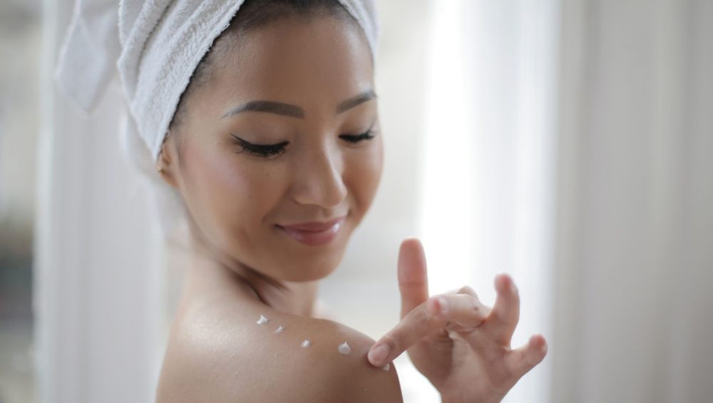 A woman with a towel wrapped around her hair dotting body lotion along her bare shoulder in soft window light