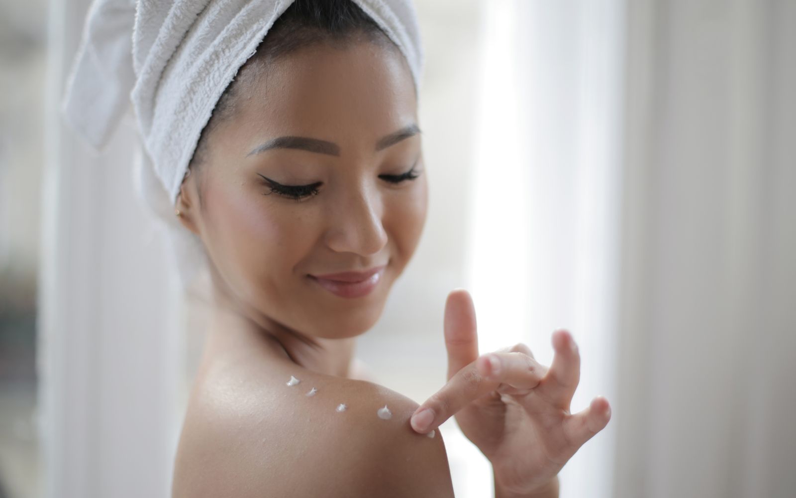 A woman with a towel wrapped around her hair dotting body lotion along her bare shoulder in soft window light