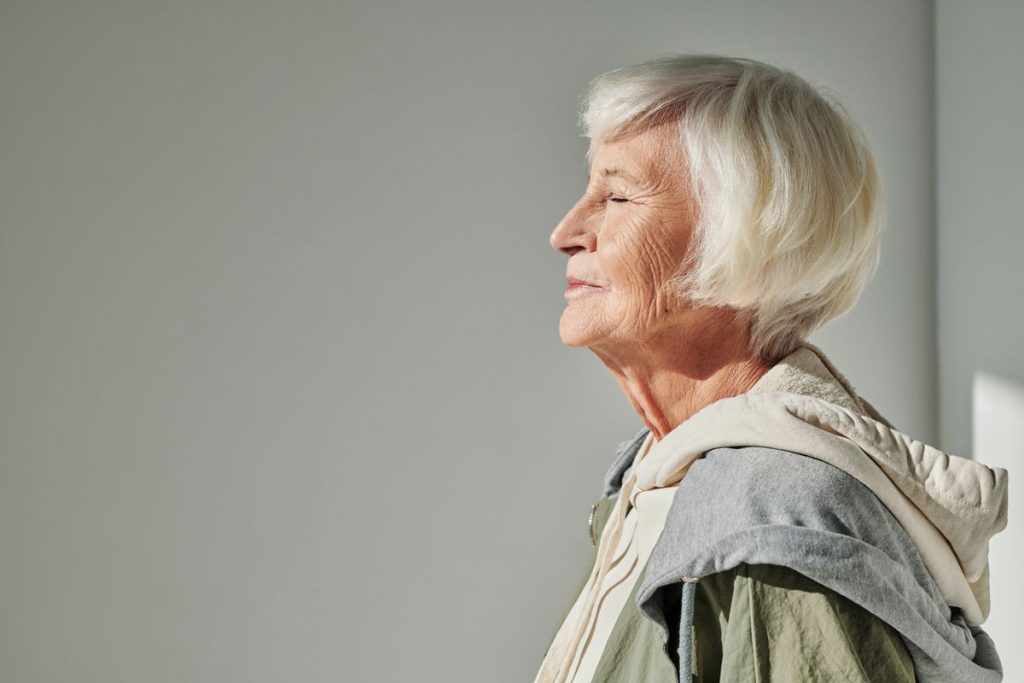 An older woman with white hair in profile in soft window light, eyes closed, no makeup