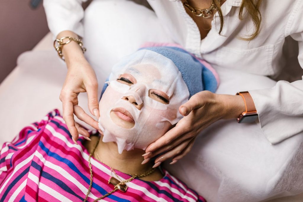 An aesthetician smoothing a white sheet mask over the face of a client lying back on a treatment bed