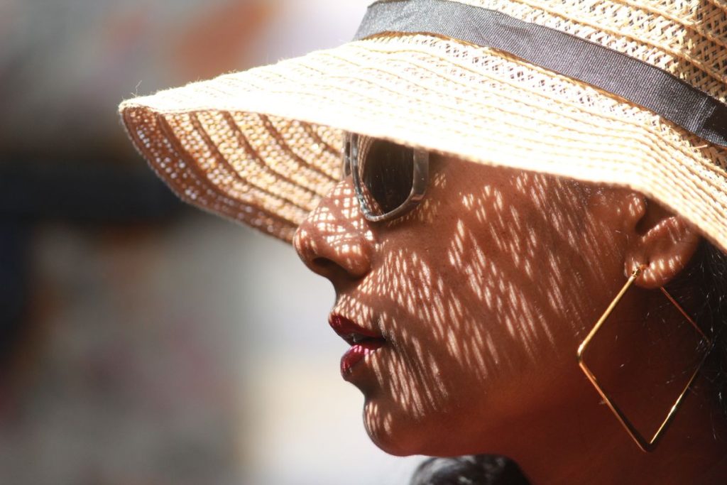A woman in a straw sunhat and sunglasses, dappled shade falling across her face