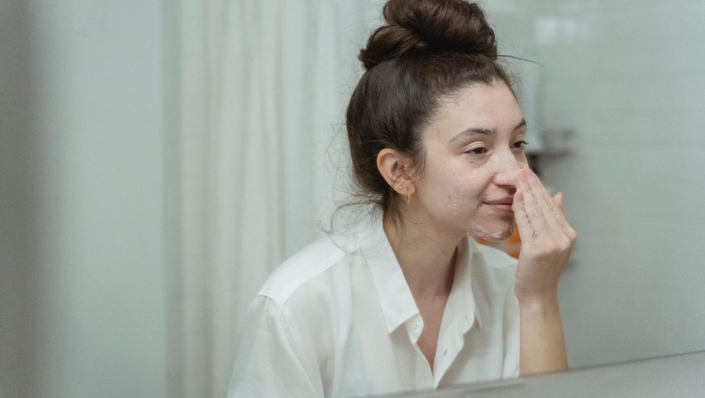 A woman in a white shirt studying her bare face in a bathroom mirror