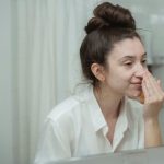 A woman in a white shirt studying her bare face in a bathroom mirror