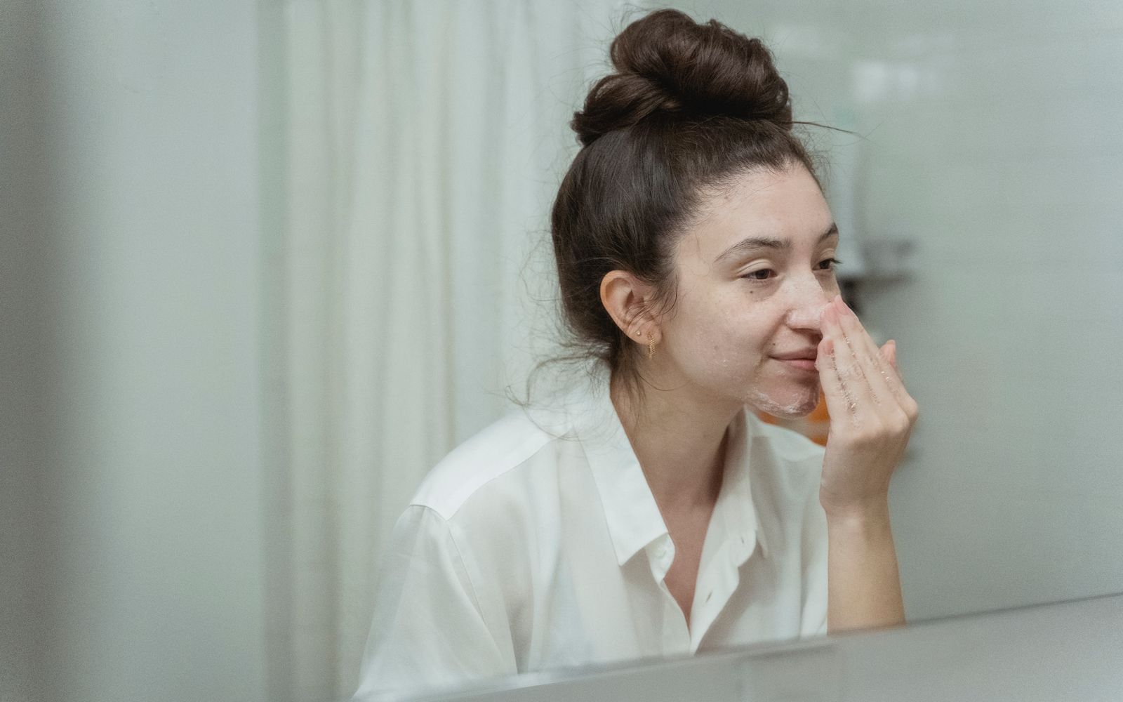 A woman in a white shirt studying her bare face in a bathroom mirror