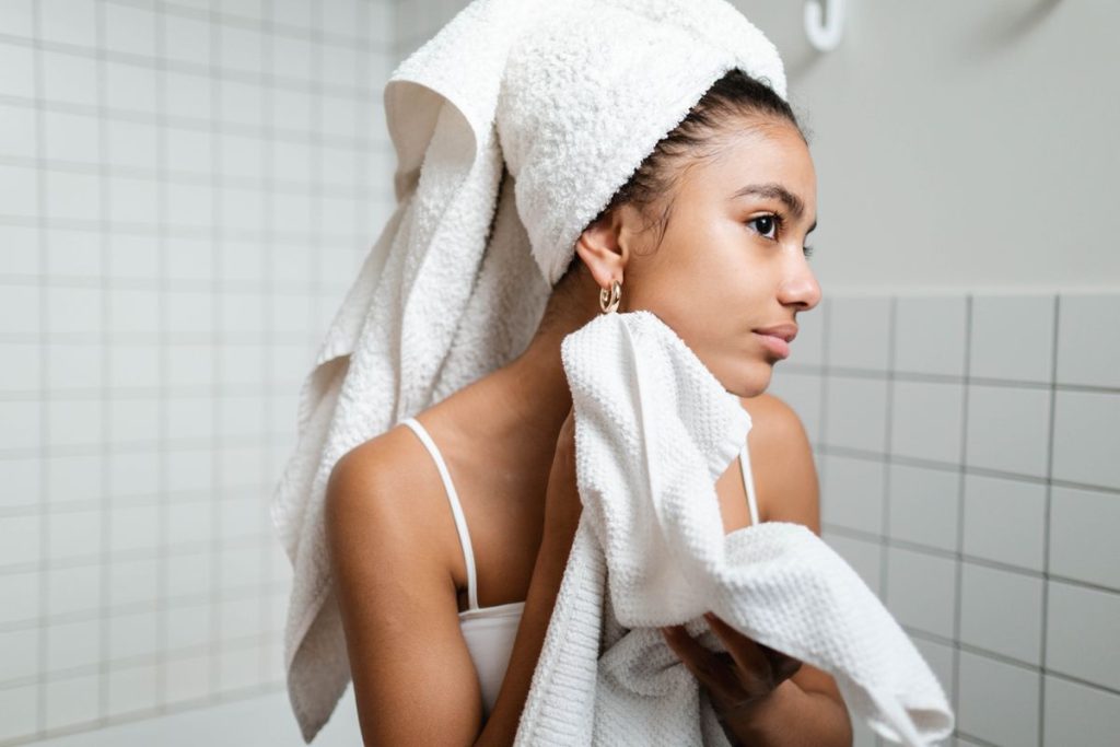 A woman with her hair wrapped in a towel pressing a soft white towel to her freshly cleansed face in a tiled bathroom