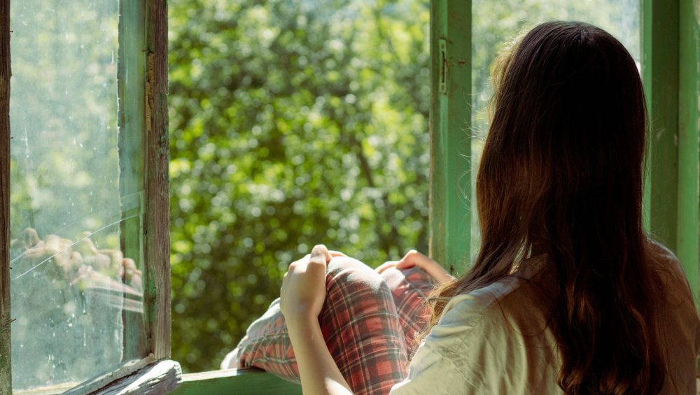 A woman sitting at a wide open window, looking out at lush green trees on a bright summer day