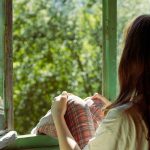 A woman sitting at a wide open window, looking out at lush green trees on a bright summer day