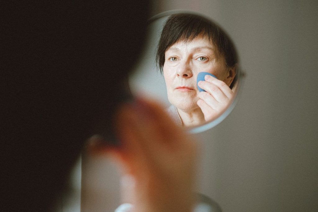 A woman in her fifties seen in a round mirror, looking closely at her face as she dabs her cheek with a soft blending sponge