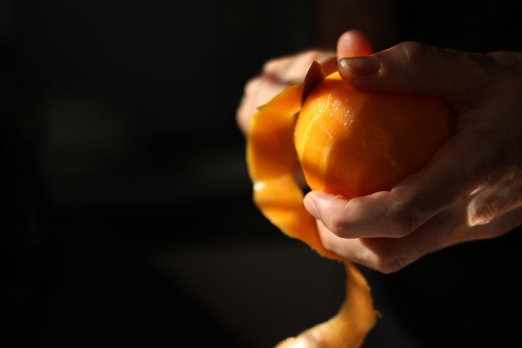 Hands peeling an orange in warm side light, a ribbon of peel falling away