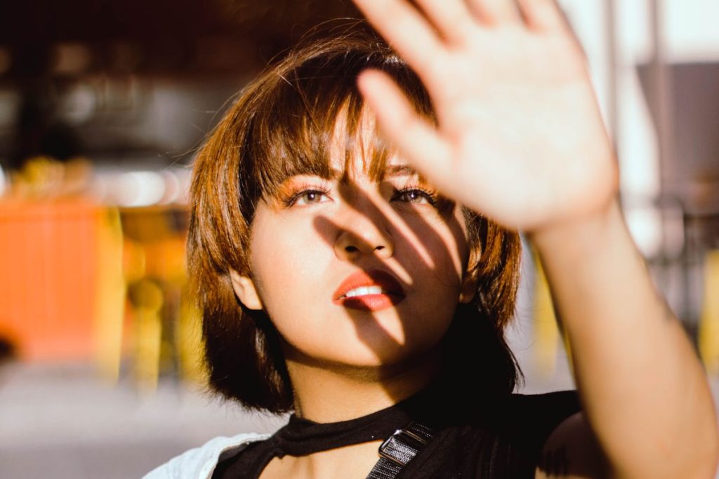 A woman outdoors in bright sun raising one hand to shade her face, light falling across her cheek and neck