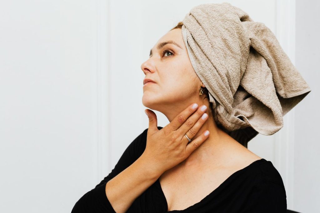 A woman with a towel wrapped around her hair smoothing her fingertips along the side of her neck in soft daylight