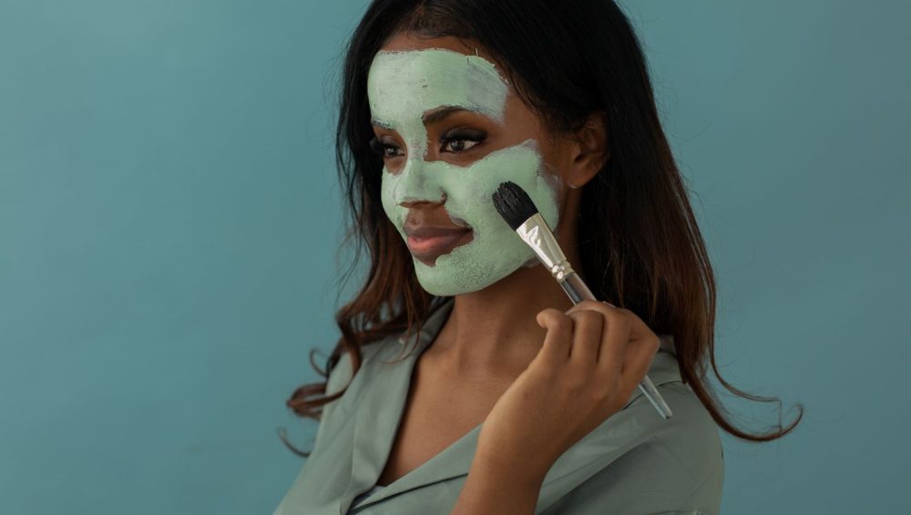 A woman smoothing a mask onto her cheek with a flat mask brush against a plain blue background