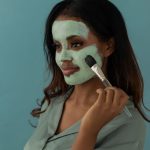 A woman smoothing a mask onto her cheek with a flat mask brush against a plain blue background
