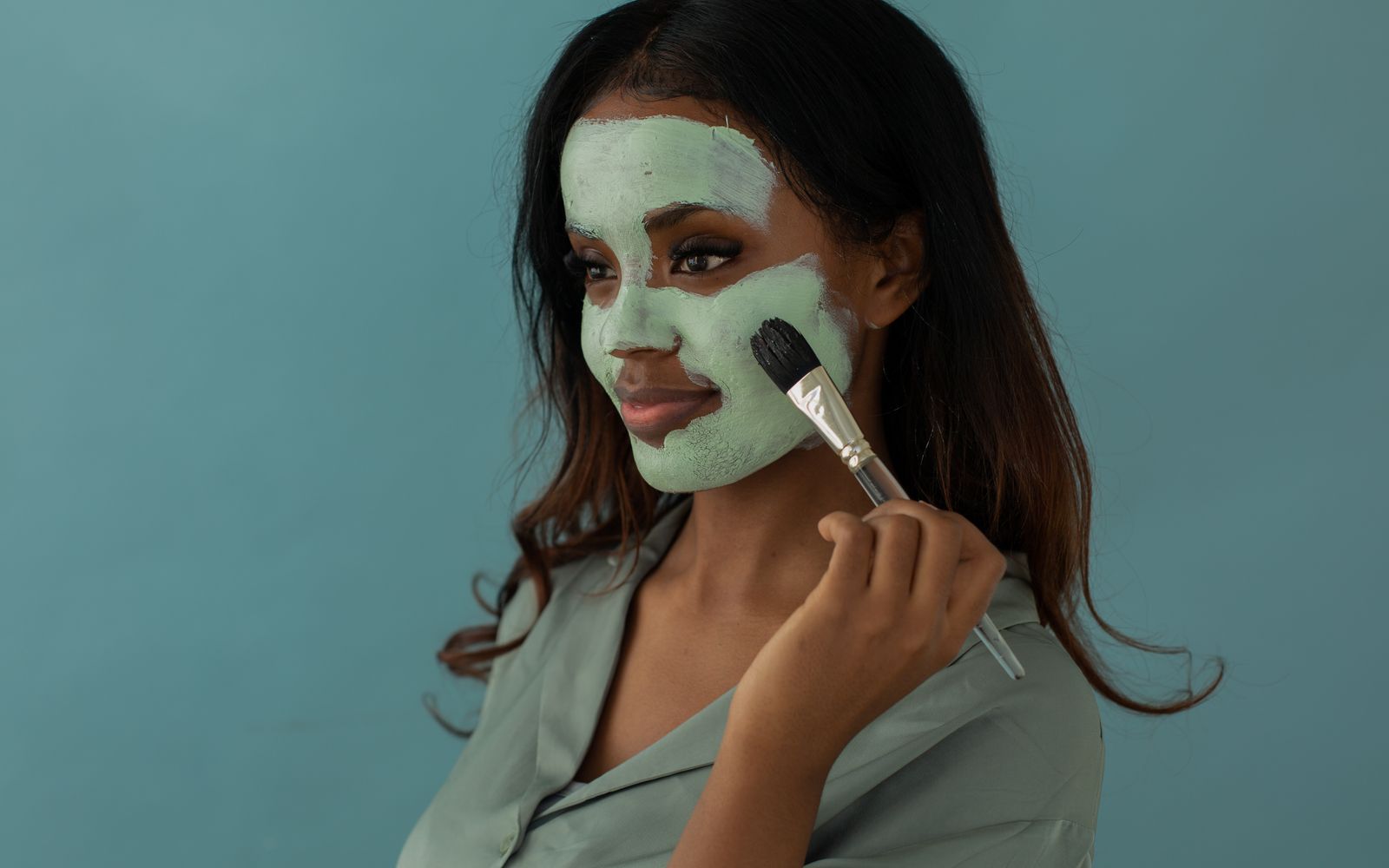 A woman smoothing a mask onto her cheek with a flat mask brush against a plain blue background