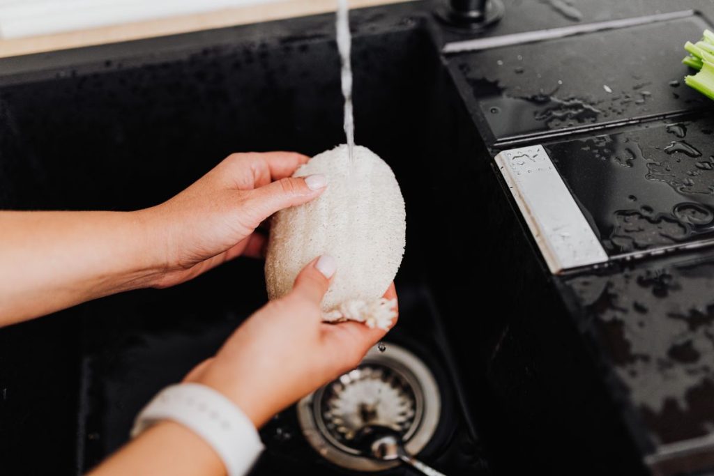 Close view of hands rinsing a soft natural sponge under a running tap at a dark sink
