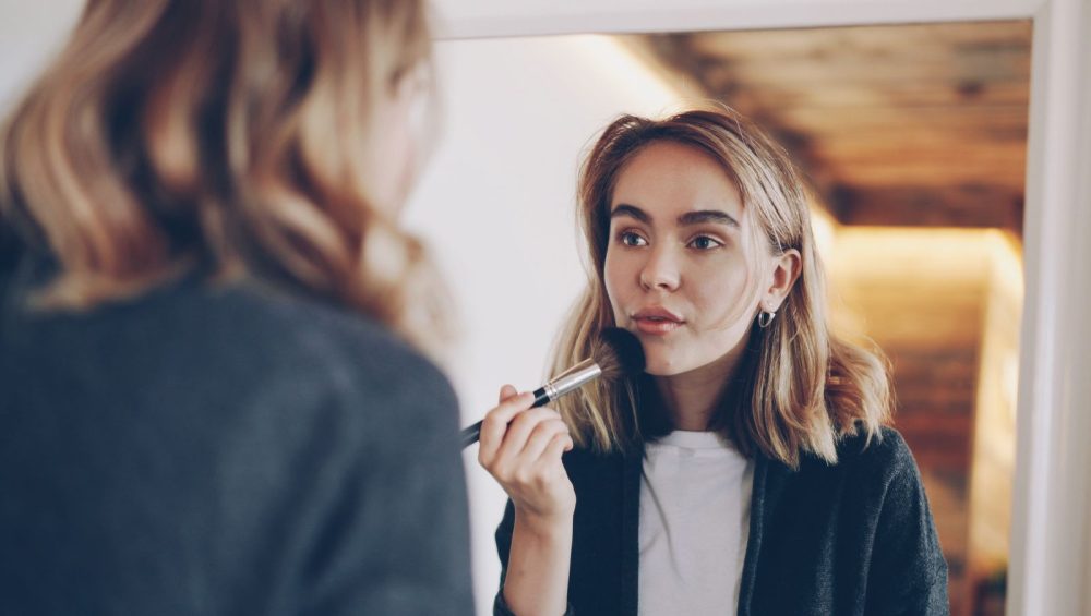 A woman at a mirror at home sweeping a soft makeup brush across her cheek