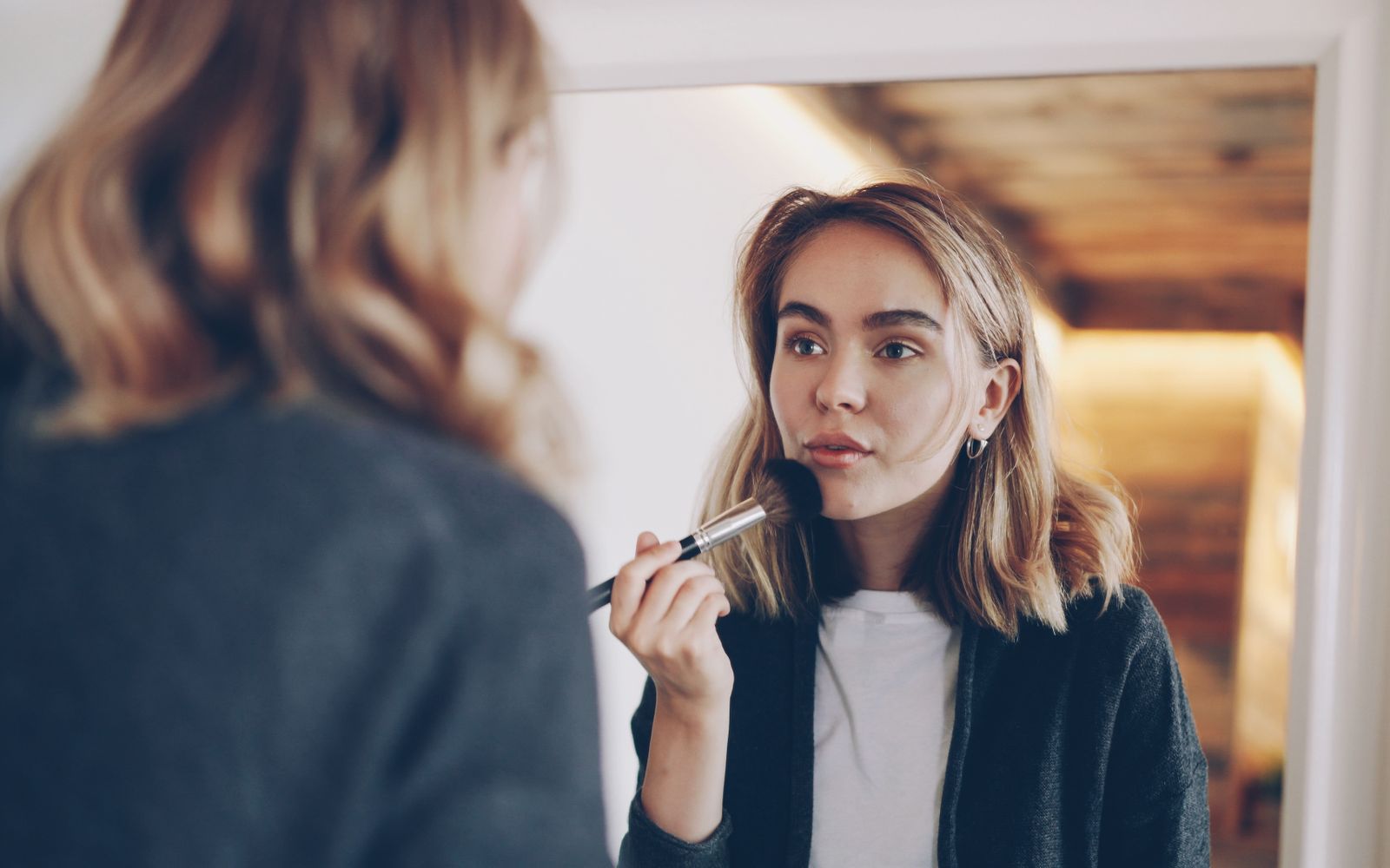 A woman at a mirror at home sweeping a soft makeup brush across her cheek
