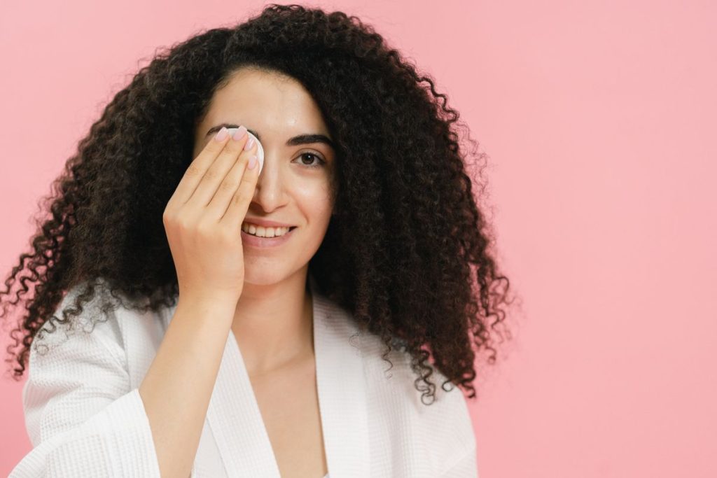 A woman in a white robe holding a soft cotton pad over one closed eye, plain pink background