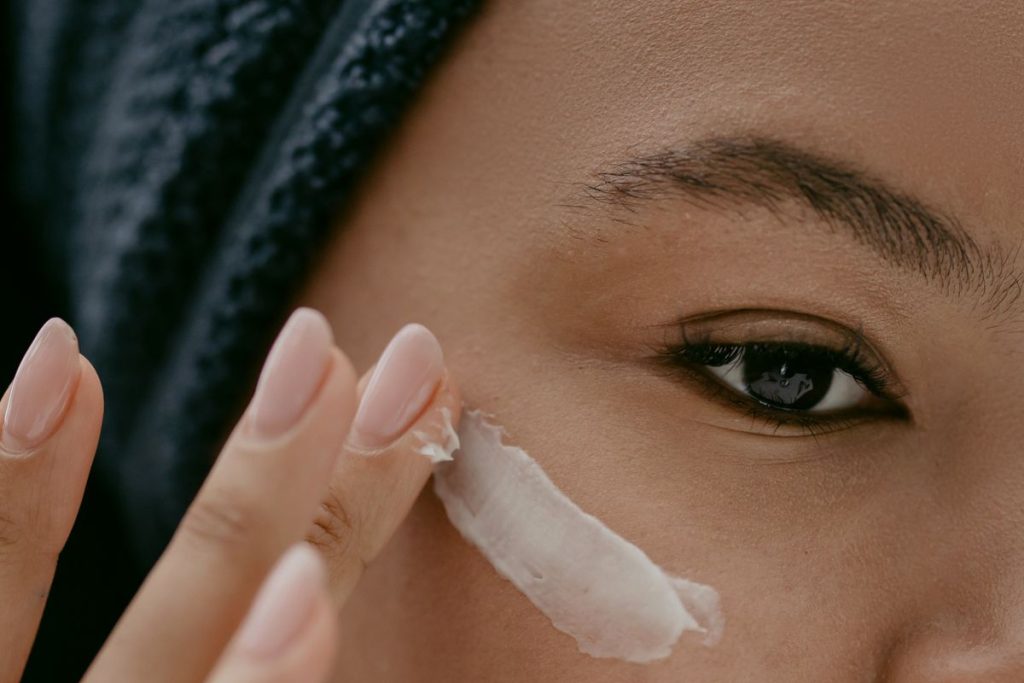 Close-up of a fingertip smoothing a small amount of white moisturizer onto a woman's cheek, no container in frame