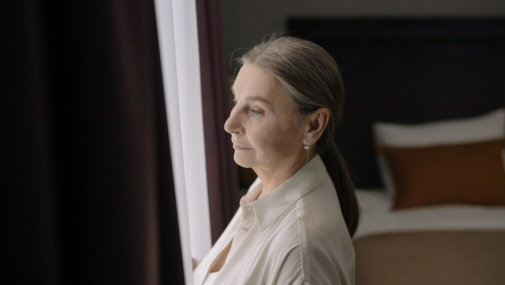 A woman in her sixties standing by a window, looking out thoughtfully
