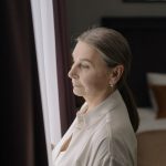 A woman in her sixties standing by a window, looking out thoughtfully
