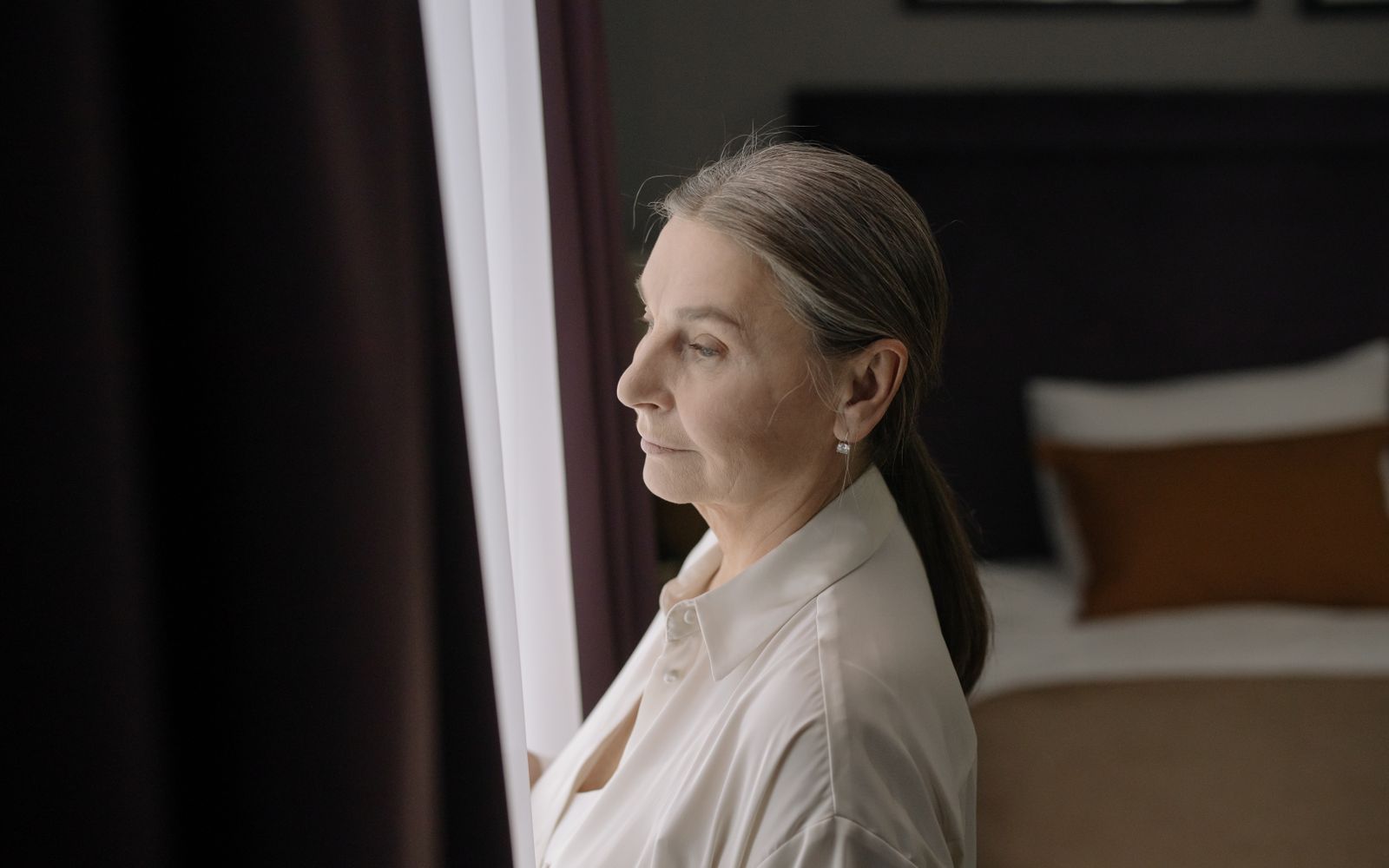 A woman in her sixties standing by a window, looking out thoughtfully