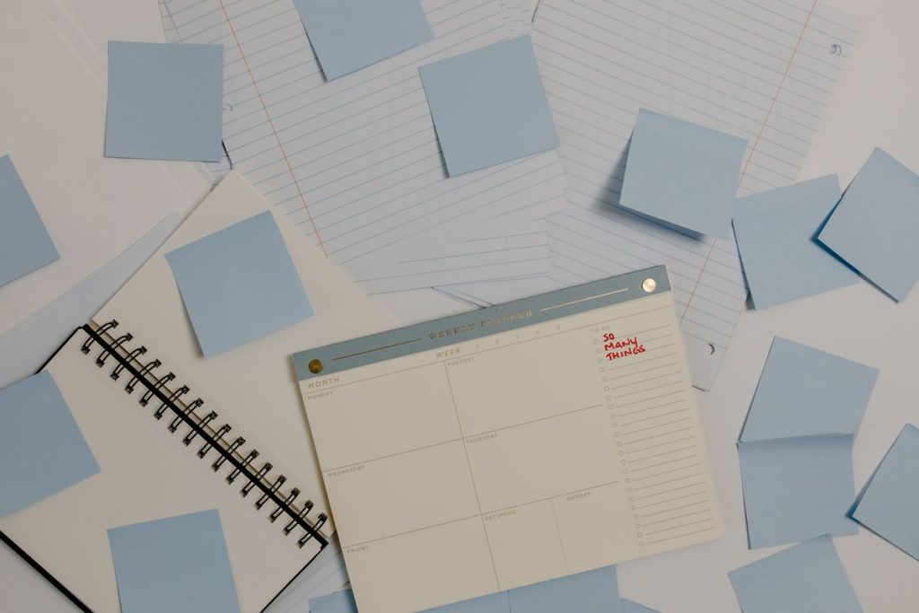 A weekly planner page lying among scattered pale blue sticky notes and a spiral notebook, one short handwritten note in the corner