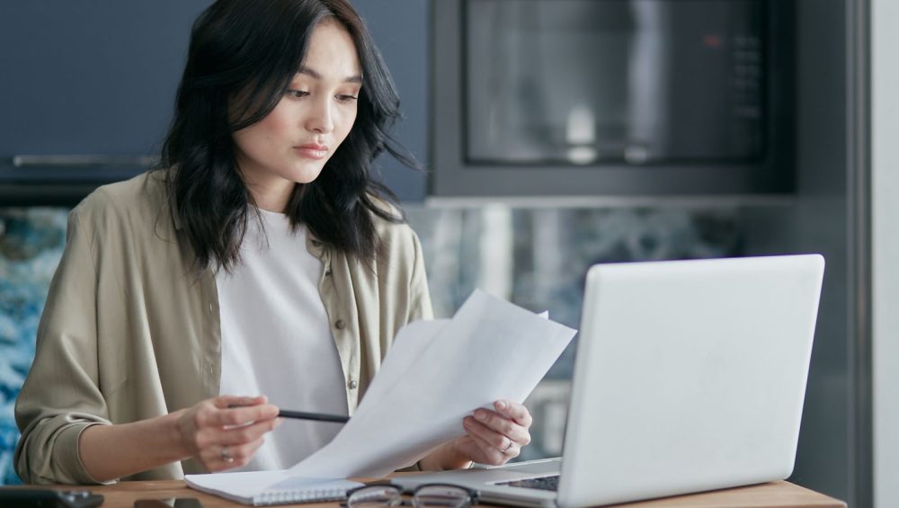 A woman at a kitchen table reading through a few printed pages, pen in hand and a laptop open beside her