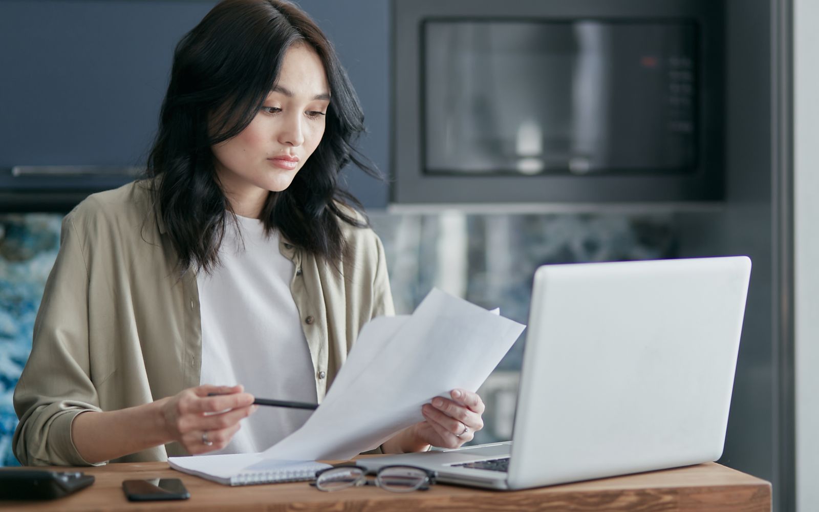 A woman at a kitchen table reading through a few printed pages, pen in hand and a laptop open beside her