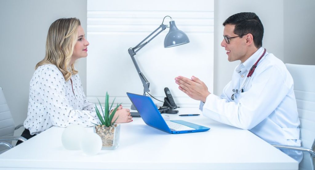 A woman seated across a white desk from a clinician in a bright consultation room, the two of them talking