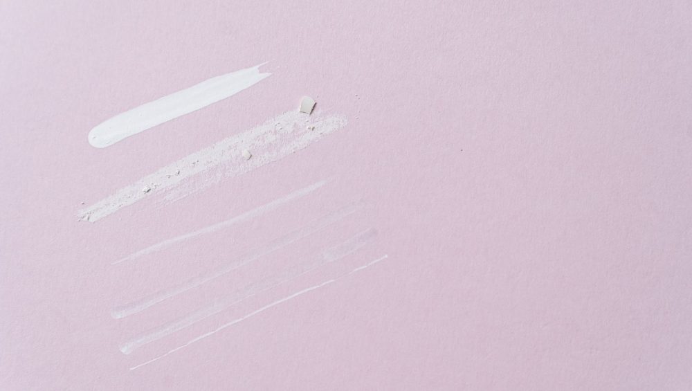 A few swatches of different textures side by side on a pale pink surface, from a thick white cream to thin, sheer strokes