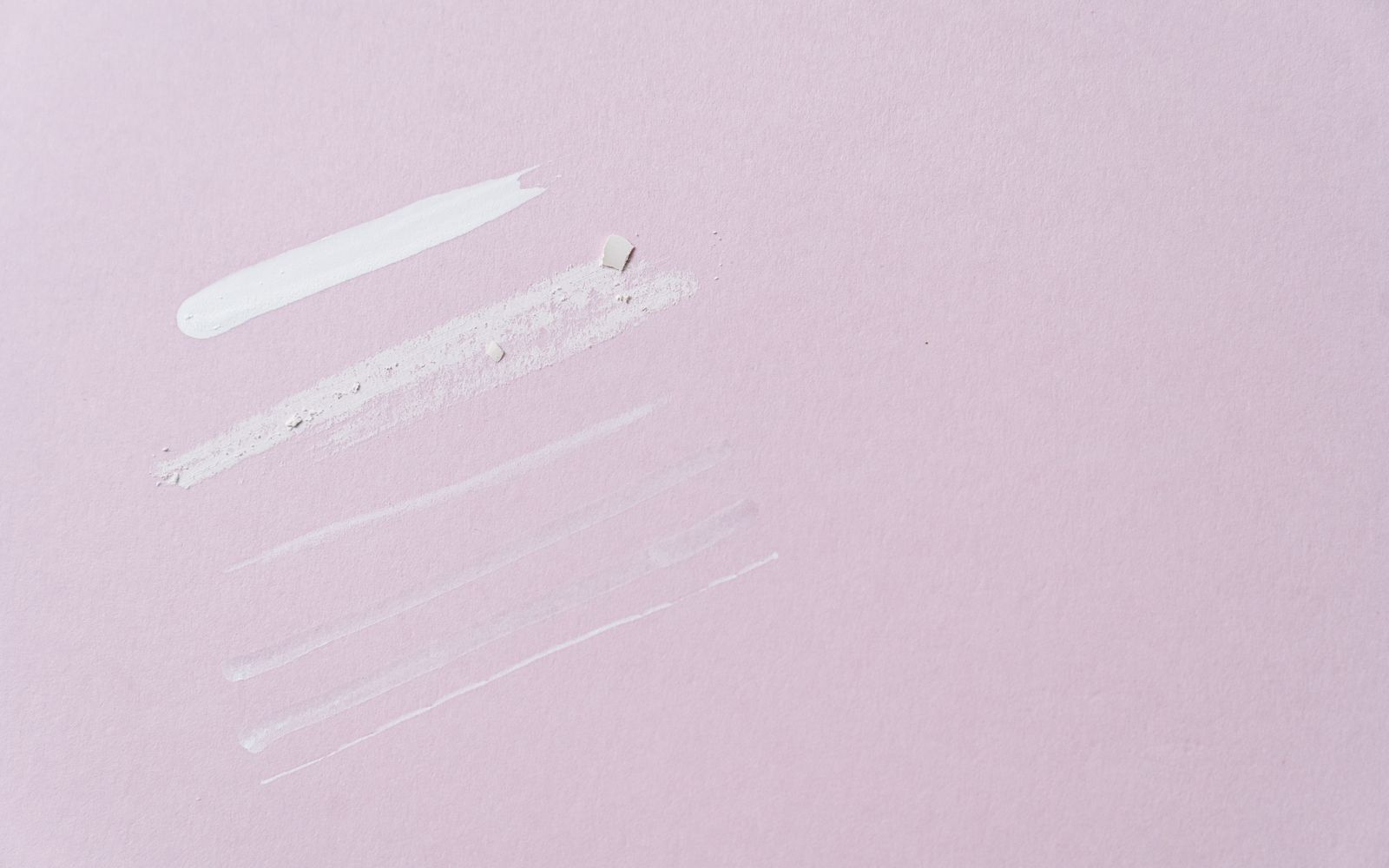 A few swatches of different textures side by side on a pale pink surface, from a thick white cream to thin, sheer strokes