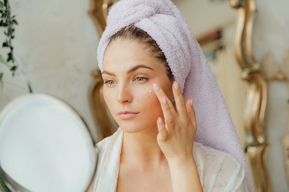 A woman with a towel wrapped around her hair smoothing moisturizer onto her cheek with her fingertips beside a mirror, soft daylight