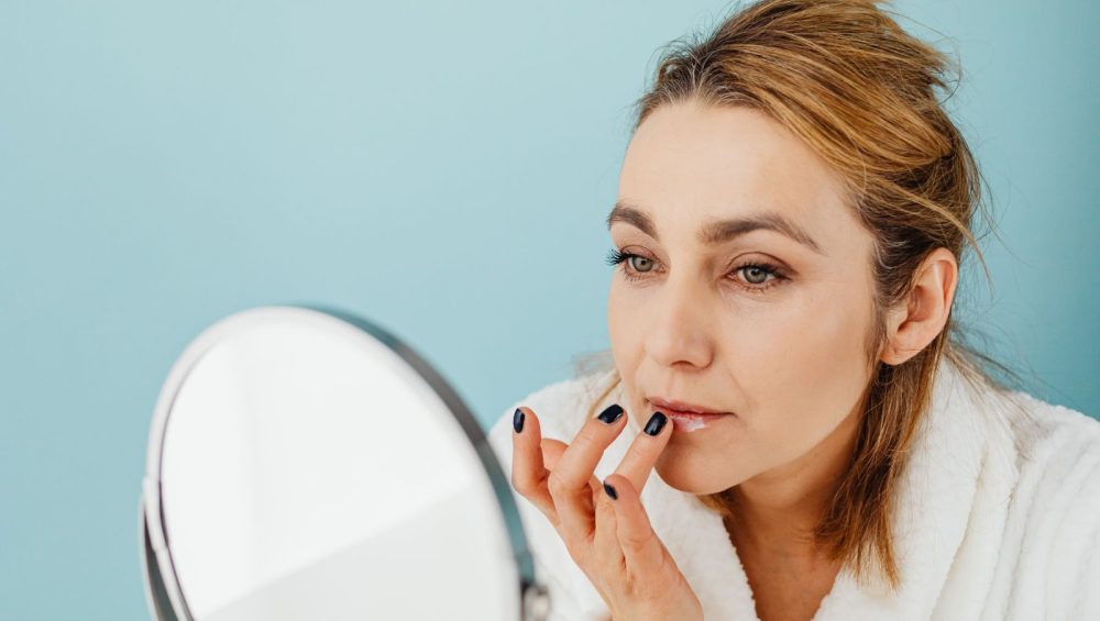 A woman in a white bathrobe at a round mirror smoothing balm onto her lips with a fingertip, bright even light