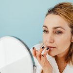 A woman in a white bathrobe at a round mirror smoothing balm onto her lips with a fingertip, bright even light