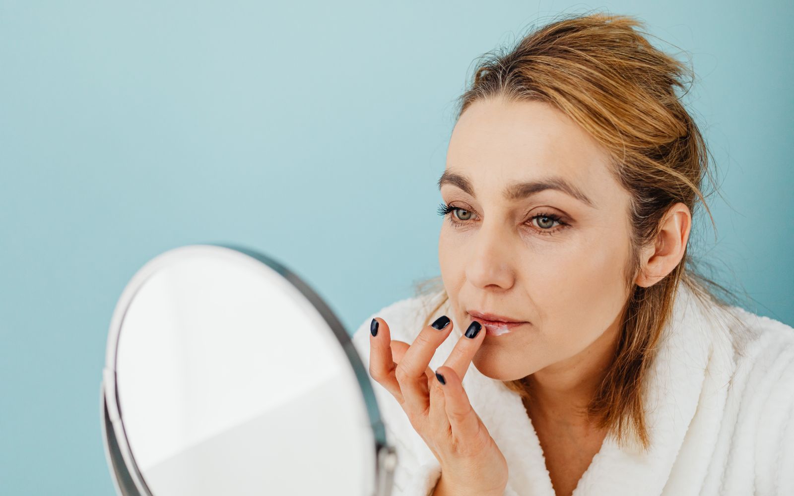 A woman in a white bathrobe at a round mirror smoothing balm onto her lips with a fingertip, bright even light
