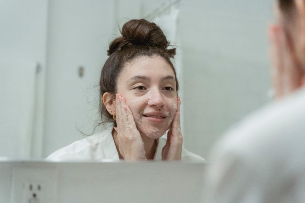 A woman at a bathroom mirror smiling as she works a light cleanser lather into her cheeks with her fingertips