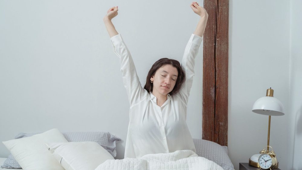 A woman stretching as she sits up in bed in early morning light