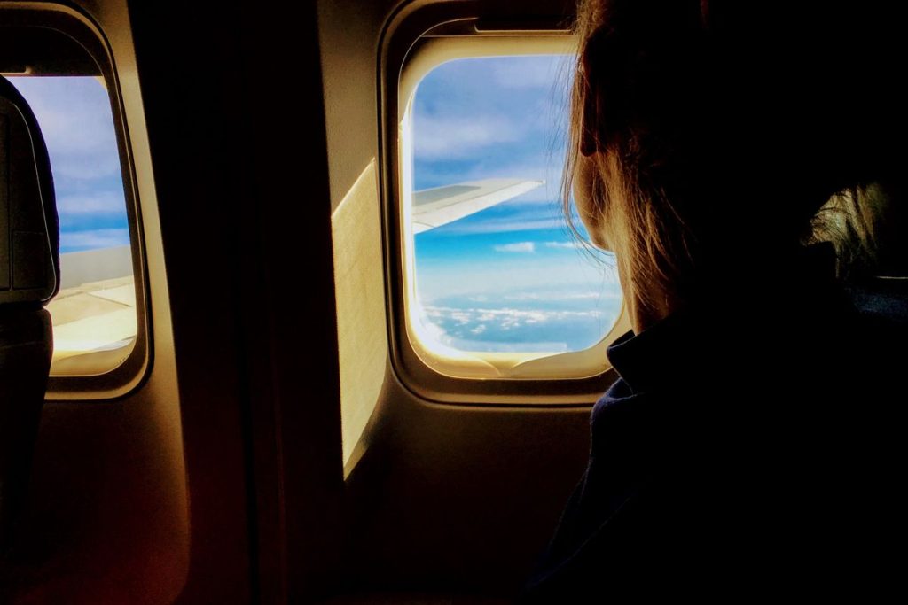 A traveler seen from behind in a window seat, the aircraft wing, blue sky and white clouds visible outside