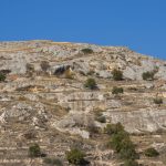 A cloudless blue sky over a dry, rocky desert hillside with sparse shrubs in strong midday light