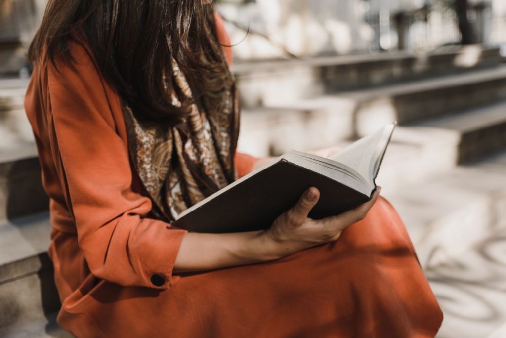 A woman in an orange coat reading a book on sunlit outdoor stone steps, her face out of frame