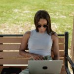 A woman in sunglasses working on a laptop on a wooden bench outdoors in bright sun
