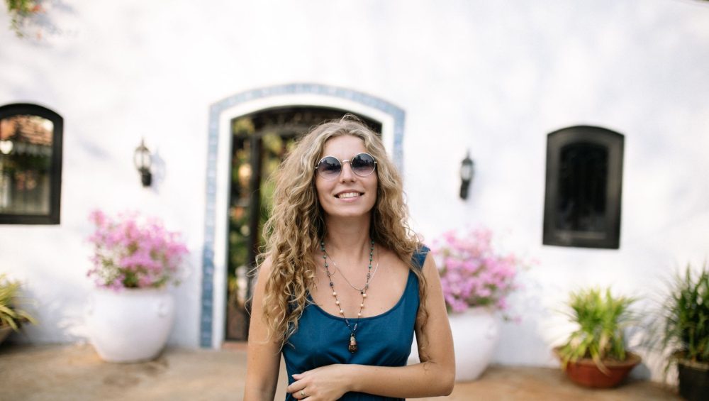 A smiling woman in sunglasses standing outside a white house with potted flowers on a bright day