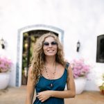 A smiling woman in sunglasses standing outside a white house with potted flowers on a bright day