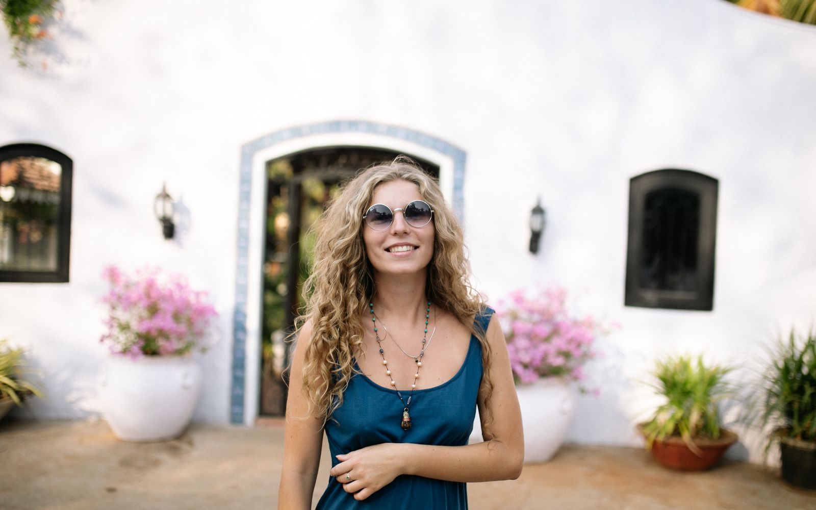 A smiling woman in sunglasses standing outside a white house with potted flowers on a bright day